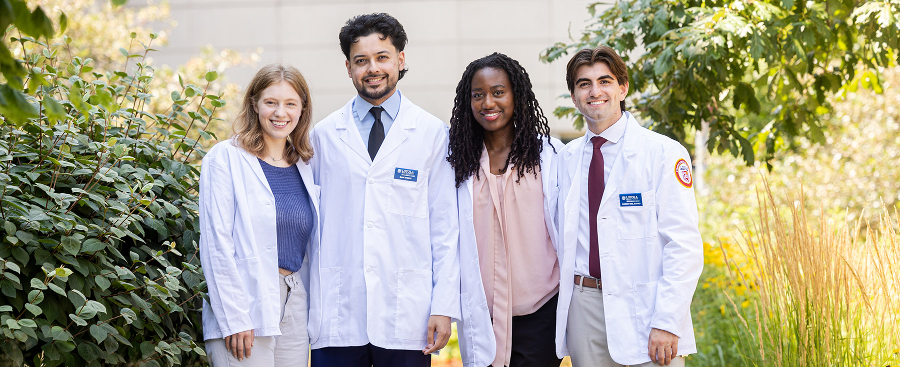 Four medical students wearing white coats smiling andstanding outside on campus 